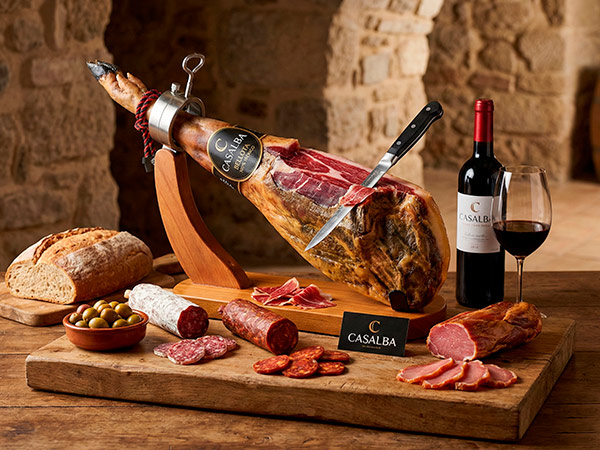 Jamón Casalba en jamonero acompañado de embutidos de diferentes tipos, aceitunas, pan y vino.