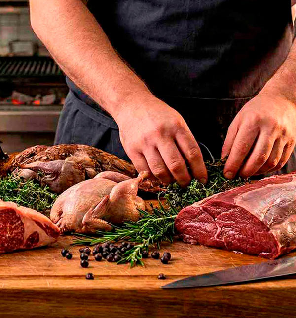 Manos de un chef profesional preparando cortes de carne roja y aves de caza selectas sobre una tabla de madera con hierbas frescas.