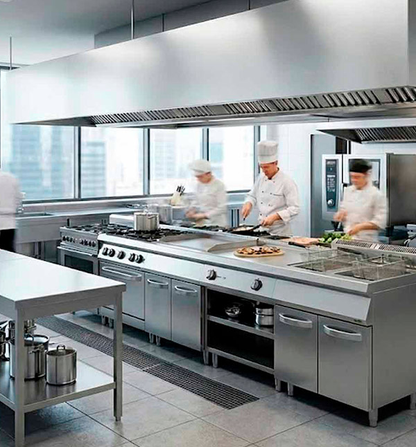 Equipo de chefs trabajando a pleno rendimiento en una cocina industrial moderna, equipada con maquinaria de hostelería de acero inoxidable, bloques de cocción y sistemas de extracción profesionales.