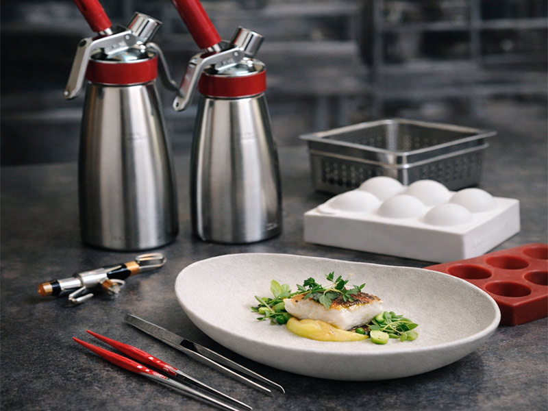 Bodegón de herramientas técnicas de cocina de Utensilios Guzmán: sifones, pinzas de precisión y moldes de silicona sobre mesa de trabajo inoxidable